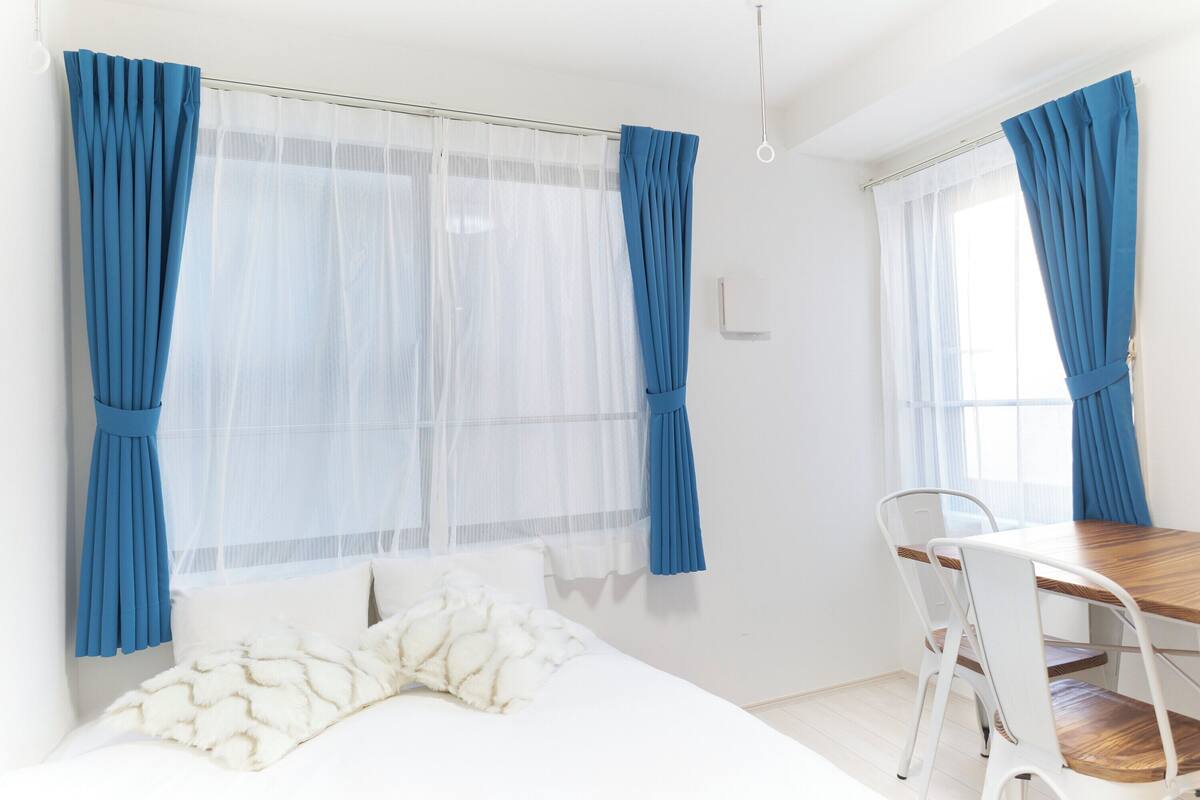 Un dormitorio con cama, una mesa de madera y sillas, y dos ventanas con cortinas azules.
