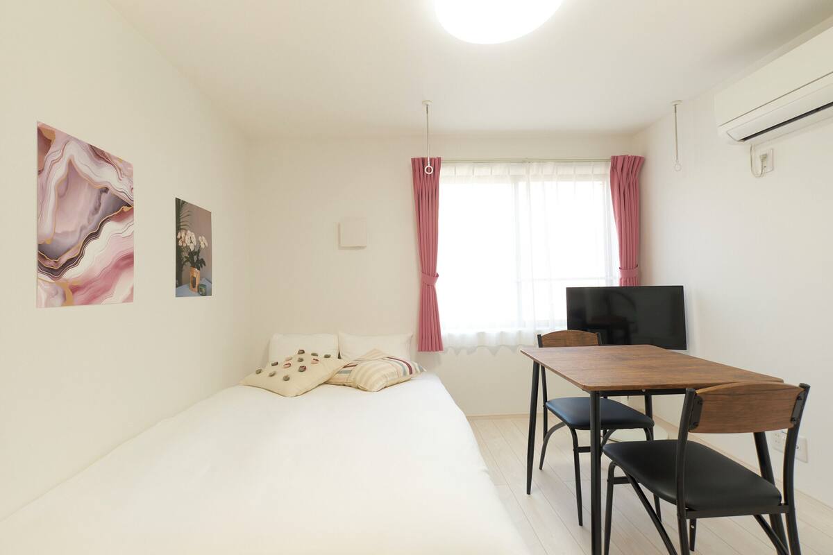 Un dormitorio con cama, escritorio y silla. Hay una ventana con cortinas, un aire acondicionado instalado en la pared y un televisor de pantalla plana.