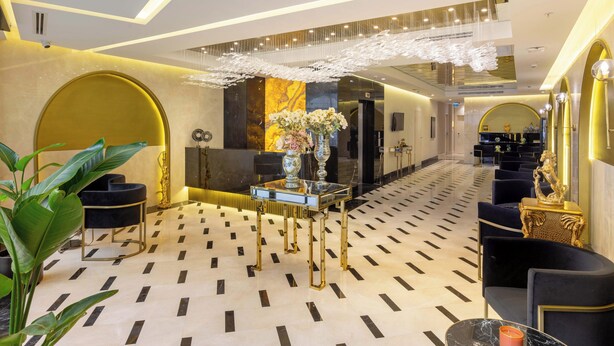 Um hall de hotel moderno com piso de mármore geométrico, balcão de recepção, área de estar e elementos decorativos como um lustre e plantas.