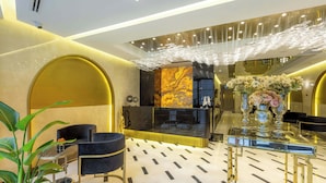 Um hall de hotel moderno, com piso de mármore, mobiliário preto e dourado, balcão de recepção e um lustre decorativo.