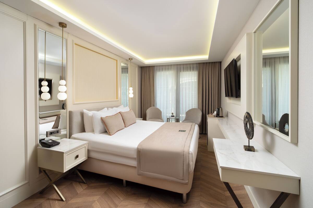 A modern hotel room with a large bed, bedside table, mirror, and a desk with a chair.