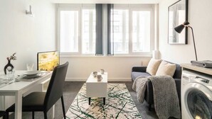 Een moderne woonkamer met een eethoek, een bank, een wasmachine en een groot raam met gordijnen.