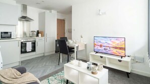Een moderne keuken met een magnetron, oven en kookplaat, een eethoek met een tafel en stoelen, en een tv op een standaard.