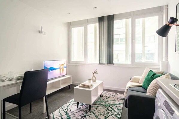 A modern living room with a flat-screen TV, a white coffee table, a black chair, a sofa with cushions, and a large window with curtains.