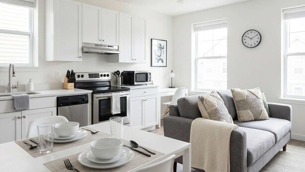 Een moderne keuken met witte kasten, een eettafel gedekt met borden, en een grijze bank in de woonkamer.