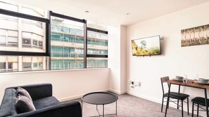 A modern living room with a sofa, a small round table, and a dining table with chairs. There is a large window with a view of a building under construction. - Central Preston Lovely 1 Bedroom Apartment (Preston)