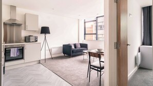 A modern kitchen with built-in appliances, a dining area with a table and chairs, a sofa, and a large window providing natural light. - Central Preston Lovely 1 Bedroom Apartment (Preston)