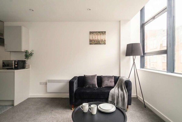 A modern living room with a black sofa, a small table with cups and plates, a floor lamp, and a window with a view. - Spacious 1 Bed Apartment in the Heart of Preston (Preston)