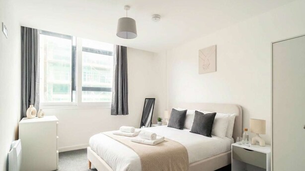 A hotel room with a bed, bedside tables, a wardrobe, a radiator, and a window with curtains. - Spacious 1 Bed Apartment in the Heart of Preston (Preston)