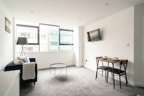A small, modern room with a sofa, a small round table, a TV mounted on the wall, and a window with a view of buildings. - Spacious 1 Bed Apartment in the Heart of Preston (Preston)