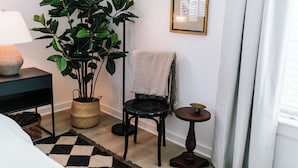 A neatly arranged room with a bed, a black chair, a small table, a lamp, a potted plant, and a framed picture on the wall. - Cozy 1BR Home by MHM Luxury Properties (Athens)