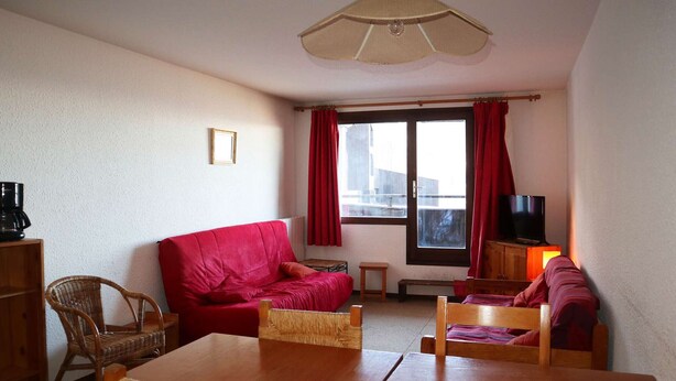 A cozy living room with a red sofa, wooden chairs, a dining table, a television, and a window with curtains.