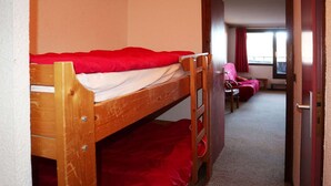 A bunk bed with red bedding in a room with a carpeted floor and a door leading to another room with a chair and a TV.
