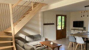 A modern living room with a wooden staircase, grey sofa, wooden coffee table, dining area with chairs, and a TV mounted on the wall. - Gîte au Cœur du Beaujolais, Dans un Domaine Viticole (buuly)
