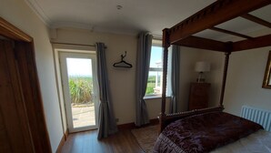 Un dormitorio con cama con dosel, suelo de madera, radiador, ventana con cortinas y vistas a la vegetación exterior.