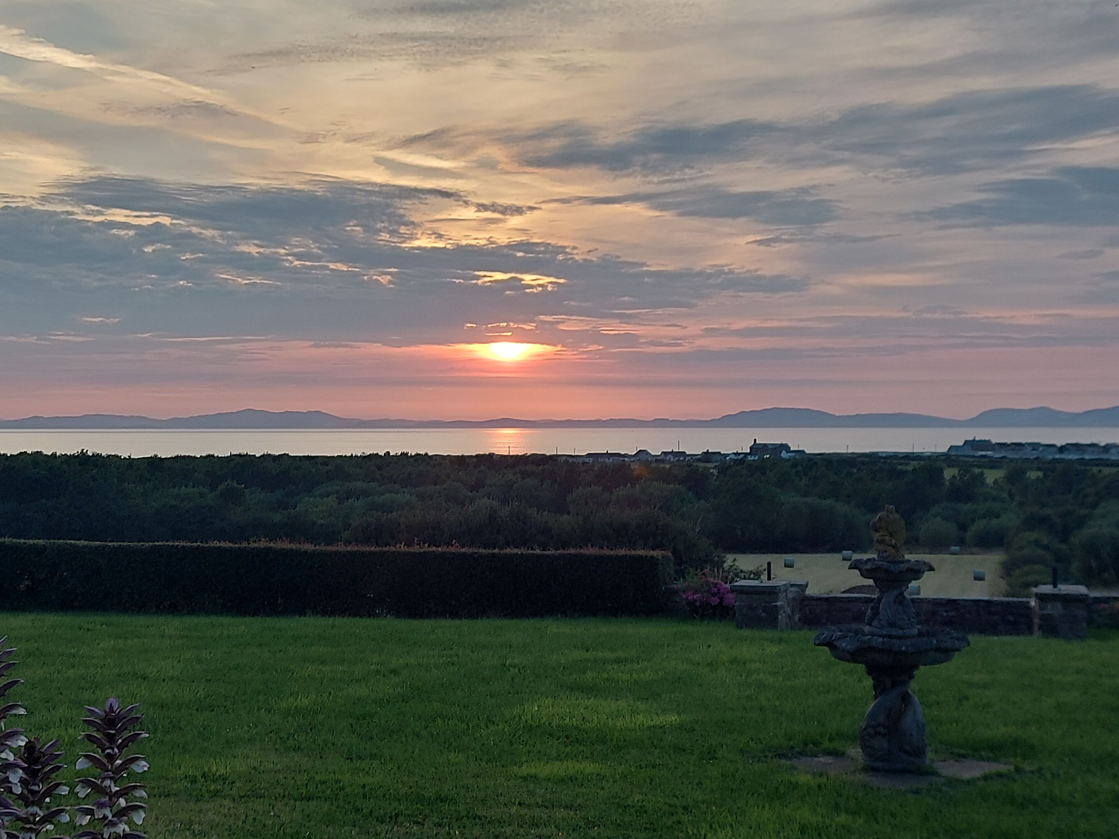 Ein Sonnenuntergang über einem Gewässer mit einer grasbewachsenen Vordergrund und einer Steinskulptur.