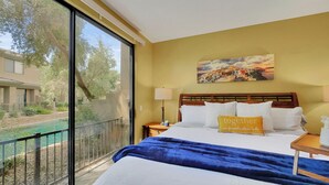 Ein Hotelzimmer mit großem Fenster, einem Bett mit weißer und blauer Bettwäsche, einem hölzernen Kopfteil und einem Gemälde einer Küstenlandschaft.