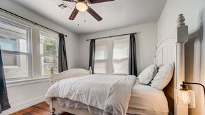 A bedroom with a bed, a ceiling fan, a bedside lamp, and a window with curtains. - 2BR Cozy House Near Paseo the Art District (Oklahoma City)