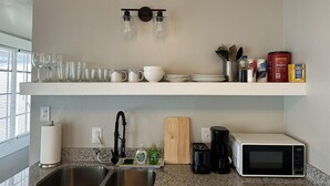 Una cucina con un piano di lavoro in granito, lavello in acciaio inossidabile, forno a microonde, tostapane e macchina da caffè. Sopra, mensole con bicchieri, piatti e utensili da cucina.