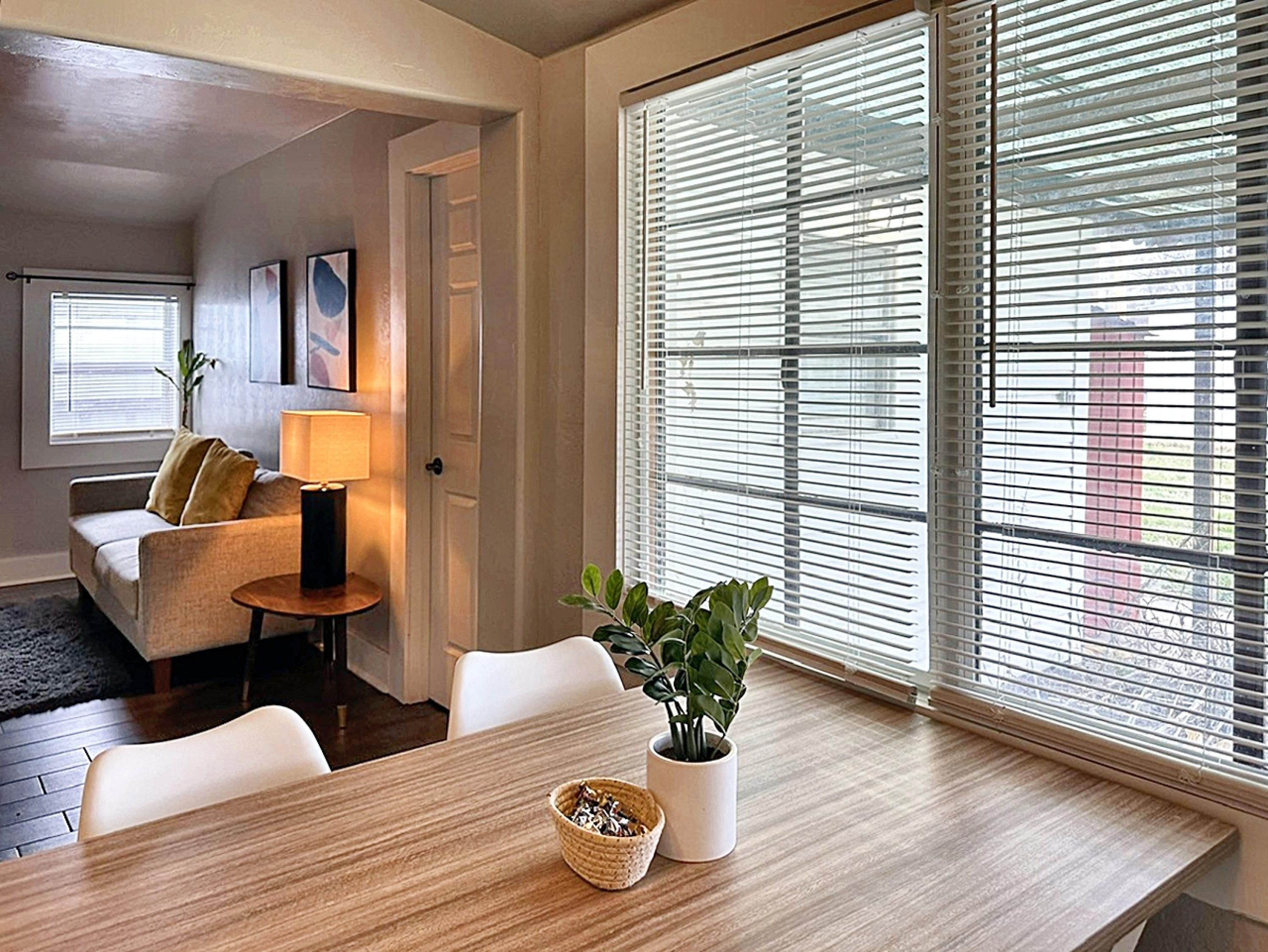 A modern living room with a wooden dining table, white chairs, a sofa with cushions, a lamp, and a potted plant.