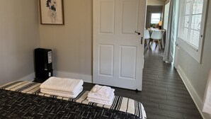 Una camera d'albergo con un letto, una porta, un termosifone, un copri-radiatore, un quadro appeso al muro, una finestra con persiane e un corridoio che conduce a una sala da pranzo.