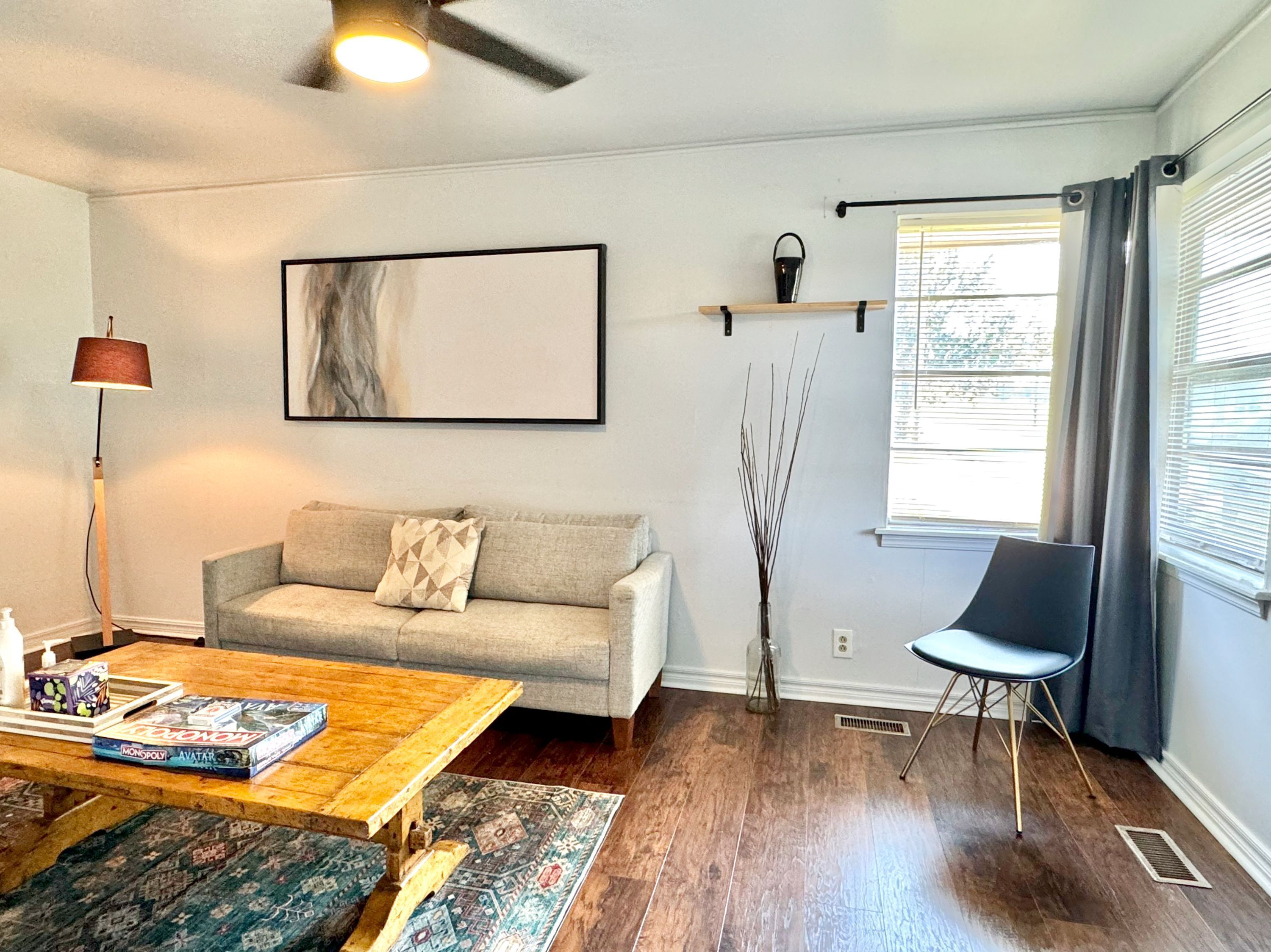 Un salon comprenant un canapé, une table basse, un fauteuil, une lampe sur pied, un tableau, un vase avec des branches et une fenêtre avec des stores.