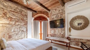 A bedroom with a brick wall, wooden ceiling beams, a bed, a bench, a TV, and a mirror. - Casa Cuero - Rooftop Pool - Historic Center (Cartagena)