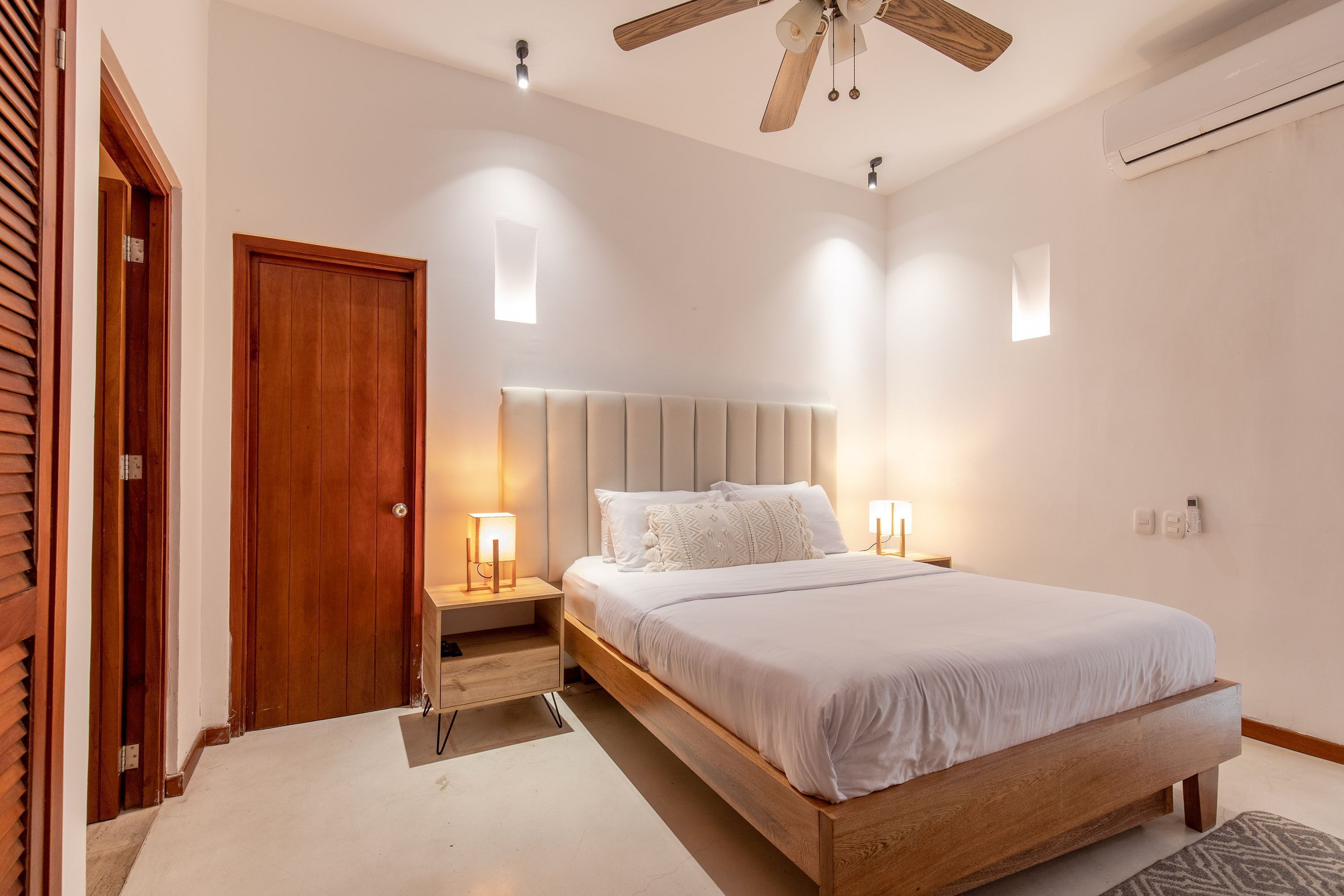A bedroom with a wooden bed, bedside lamps, a ceiling fan, and an air conditioning unit.
