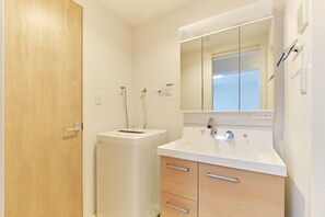 Une salle de bain avec une porte en bois, un lave-linge blanc, un lavabo avec un miroir au-dessus et un meuble avec des tiroirs.