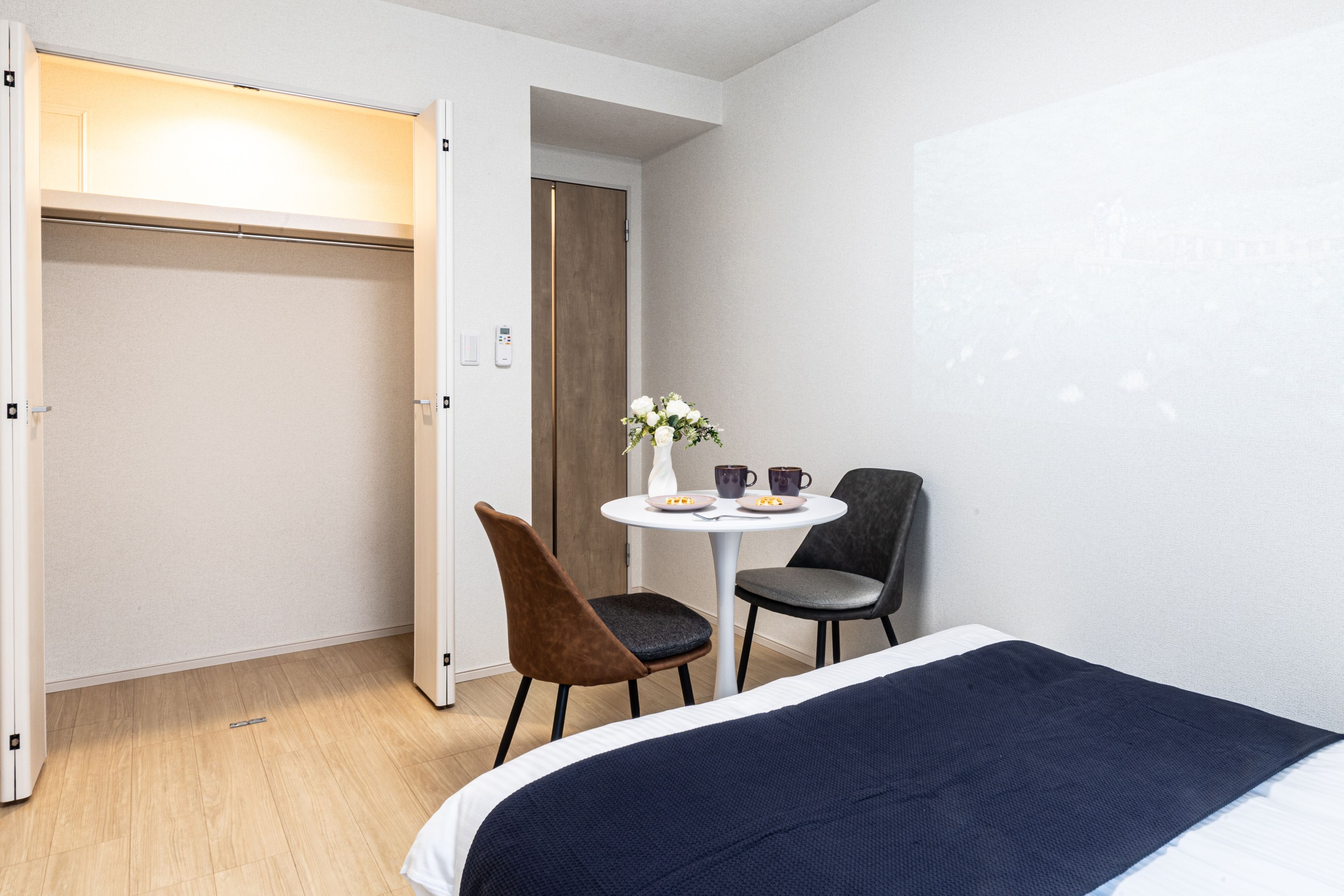 A hotel room with a bed, a desk with two chairs, a small table with a vase and cups, and a wardrobe.