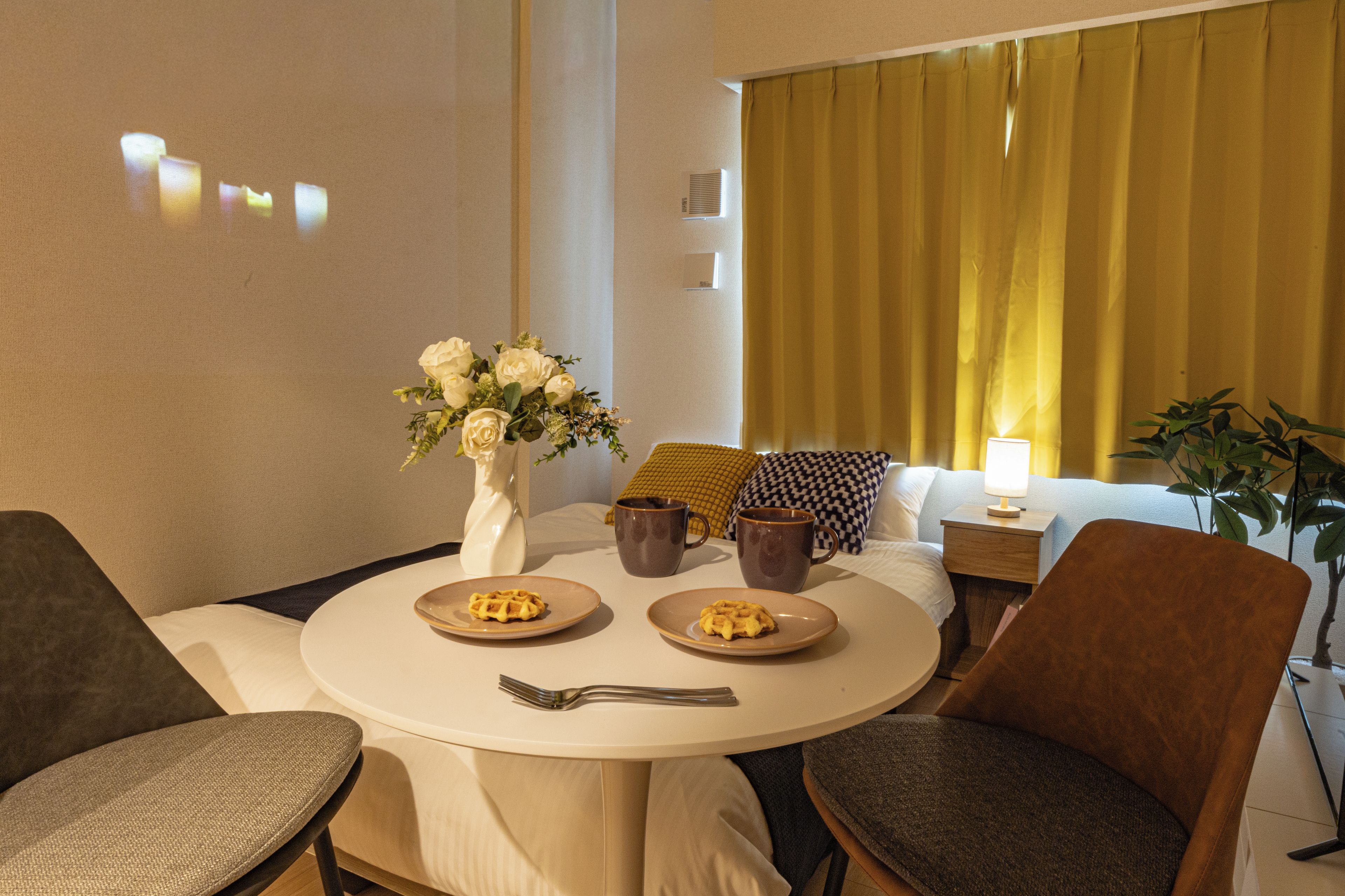 Hotellihuone, jossa on pyöreä ruokapöytä, kaksi tuolia, maljakko kukkineen, lamppu ja huonekasvi.