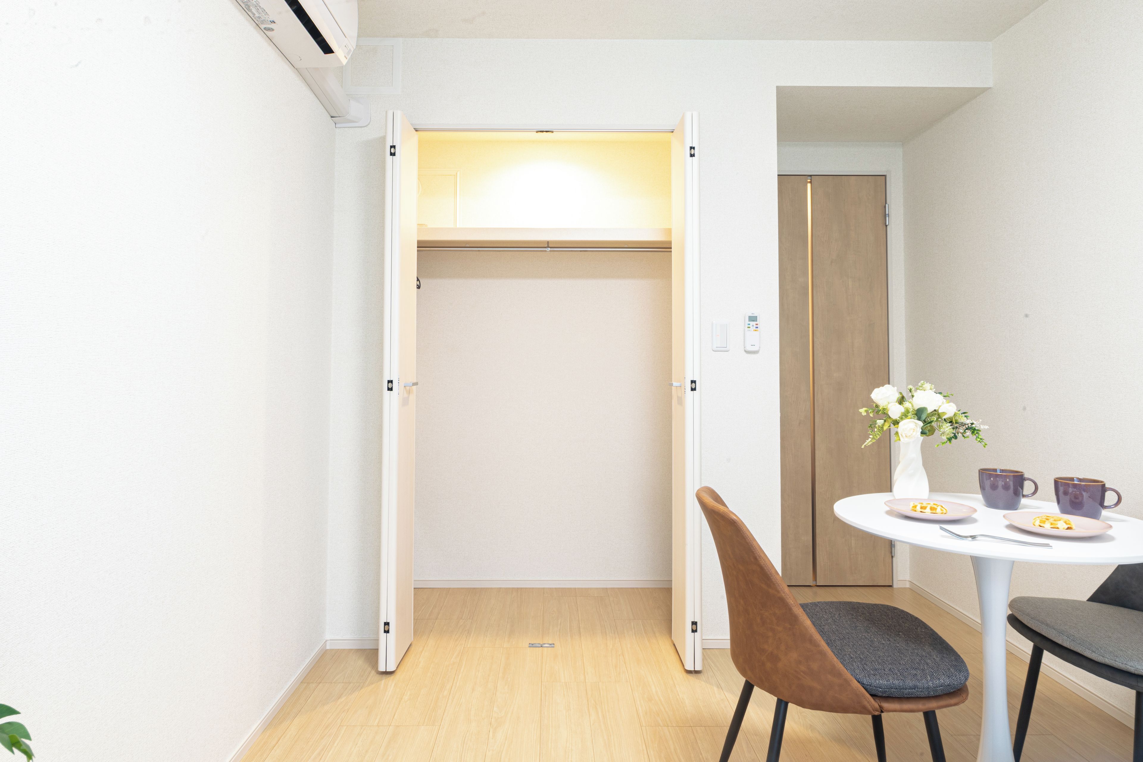 A compact room with a white wardrobe, a small dining table with two chairs, and a vase with flowers.
