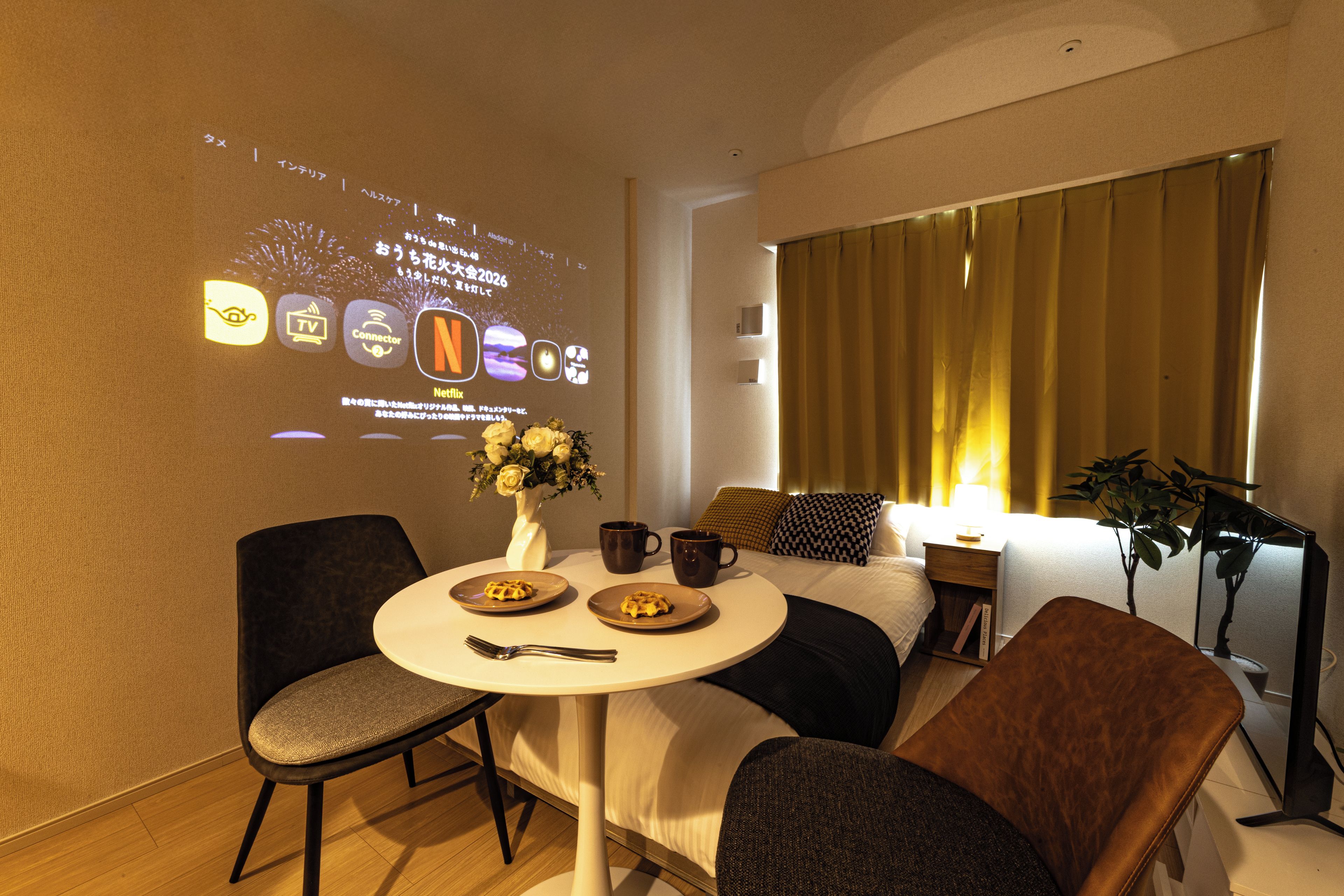 A hotel room with a bed, a bedside table with a lamp, a chair, a small table with a vase of flowers, and a wall-mounted projector displaying hotel information.