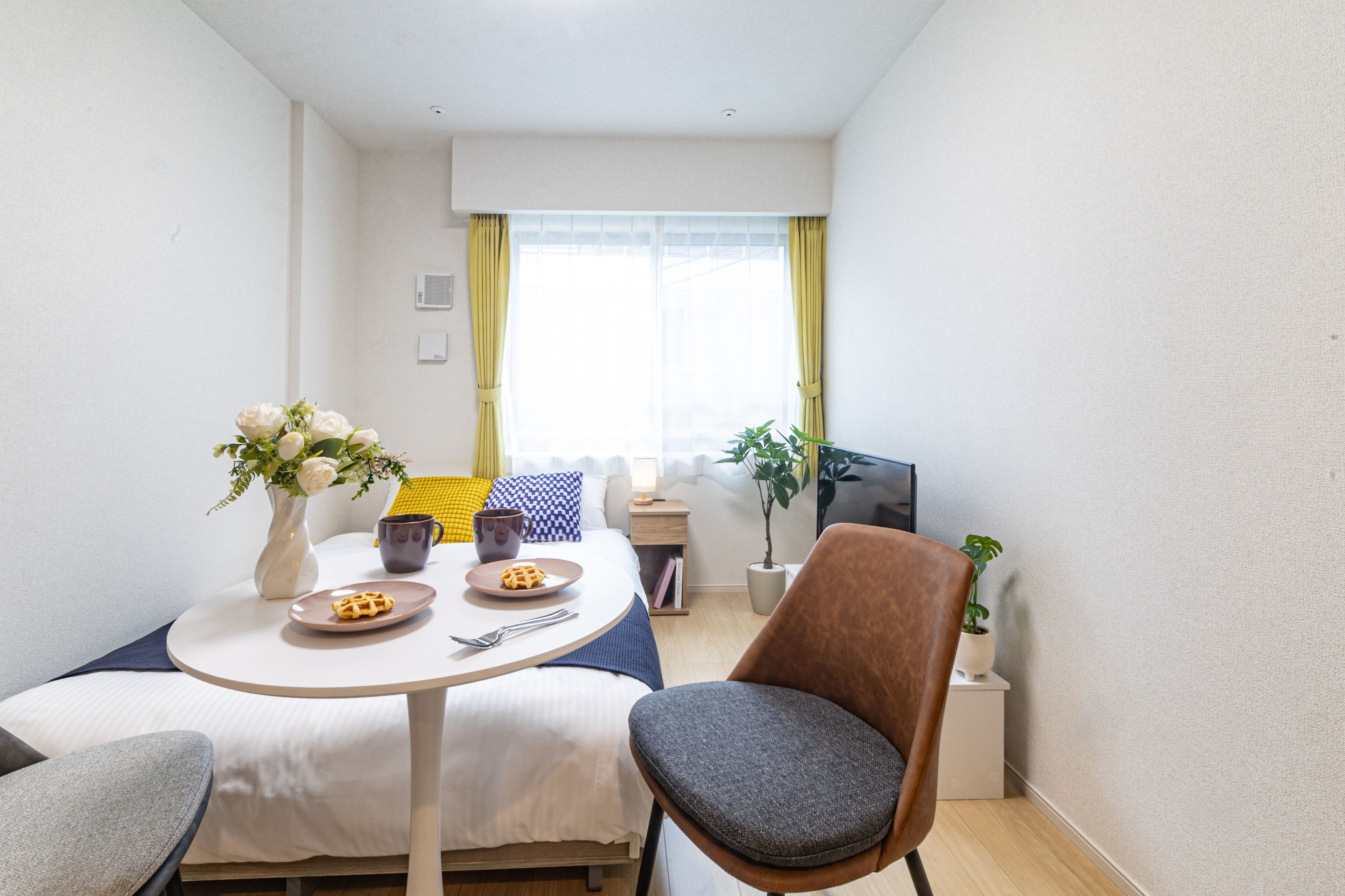 A small, well-lit room with a bed, a table with a plate of food, a chair, a TV, and a window with curtains.