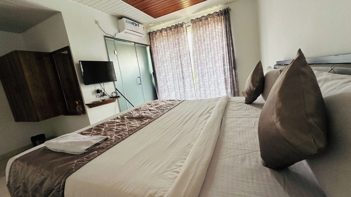 Una habitación de hotel con una cama, un televisor de pantalla plana montado en la pared, una ventana con cortinas y un ventilador de techo.