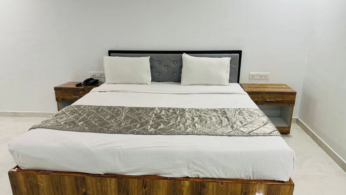 Una cama bien hecha con colcha gris, dos almohadas blancas y cabecera de madera. Hay dos mesitas de noche, una con teléfono y la otra con lámpara.