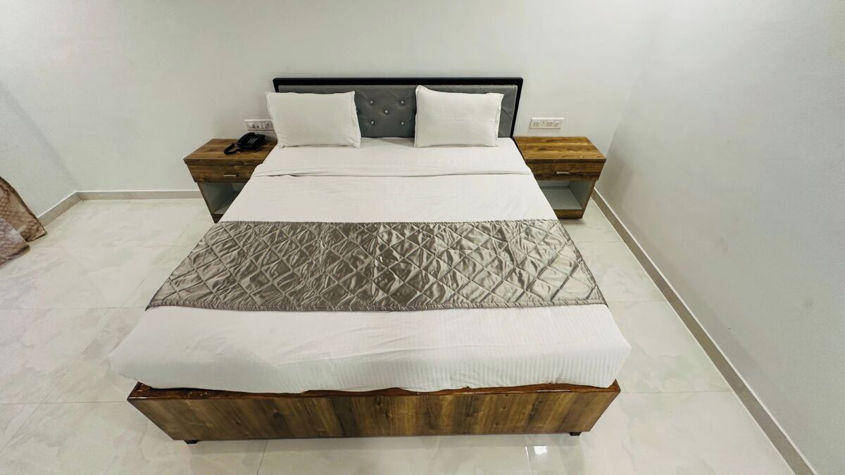 Een netjes opgemaakt bed met een gewatteerd hoofdbord, een houten nachtkastje met een telefoon, en een bijzettafeltje met een lamp.