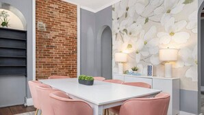 Zona de menjador amb taula blanca i cadires roses, paret de maó i paper pintat floral.