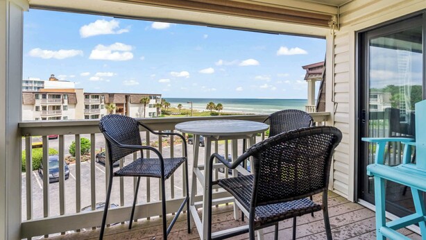 A balcony with two wicker chairs and a small table, overlooking a beach and buildings. - Ocean Forest Villas Unit D304 - Ocean view (Myrtle Beach)