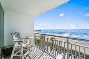 A balcony with a view of the ocean and a pier, featuring a white Adirondack chair and a small table.