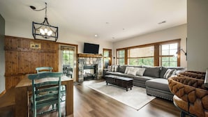 A spacious living room with a fireplace, a dining area with wooden furniture, and large windows. - Mountain Creekside: Hot Tub: Golf (Boyne City)