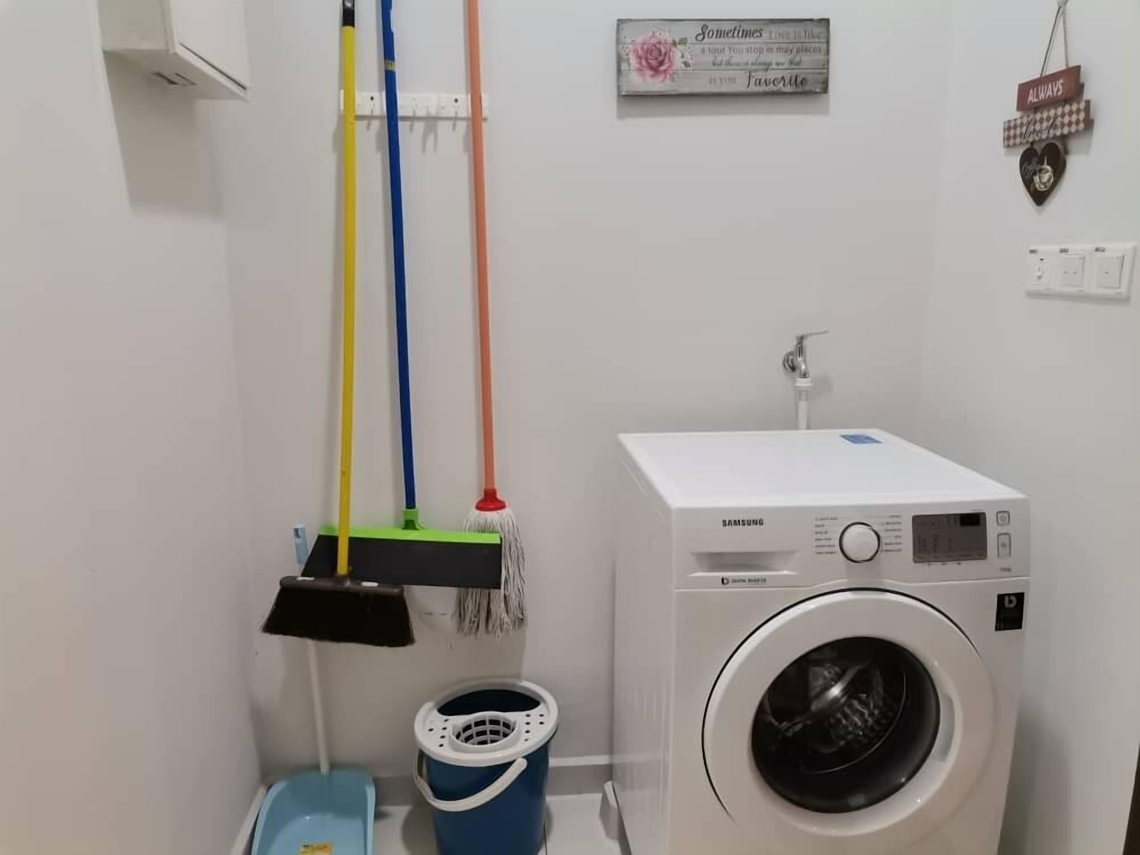 Un cuarto de lavado con una lavadora Samsung blanca, un balde azul y tres escobas colgadas en un tendedero.