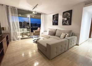 A modern living room with a sofa, a glass door leading to a balcony, and two framed artworks on the wall.