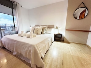 A bedroom with a large bed, a bedside table with a lamp, a mirror on the wall, and a view through a window.