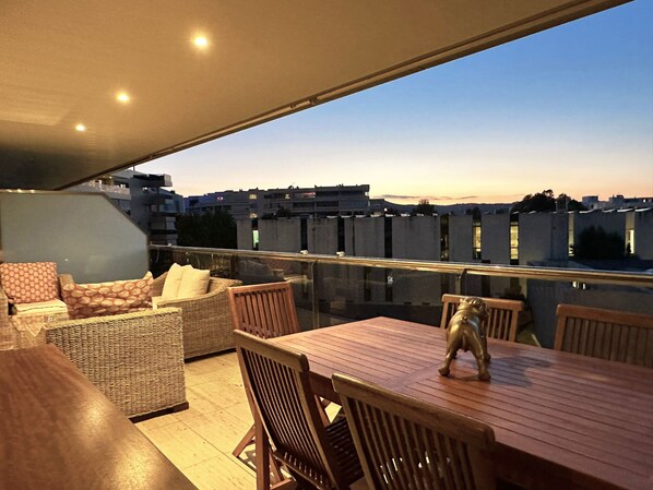 A rooftop terrace with a dining table and chairs, a wicker sofa, and a view of the cityscape at dusk.