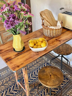 A wooden table with a vase of purple flowers, a bowl of lemons, a wicker basket, and a vintage suitcase.