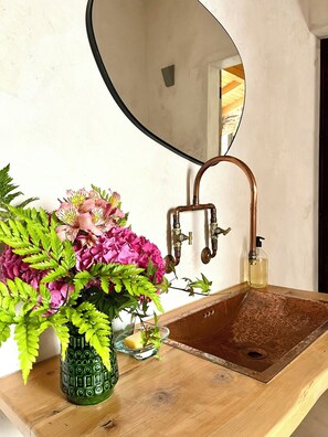 A bathroom with a wooden vanity, a copper sink, a mirror, and a vase of flowers.