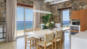 A modern kitchen with a stone wall, a dining area with a white table and woven chairs, and a view of the sea. - Stylish Villa w/Private Pool & BBQ near Elafonissi (Chania)