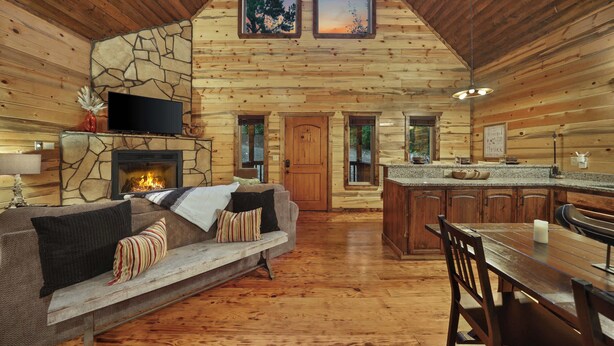 A log cabin interior with a stone fireplace, a sofa, a television, a dining table, and a kitchen area.