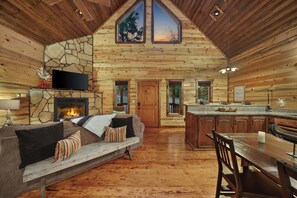 A log cabin interior with a stone fireplace, a sofa, a television, a dining table, and a kitchen area. - Rustic Cabin Near Creek with Hot Tub & Fire Pit (Broken Bow)